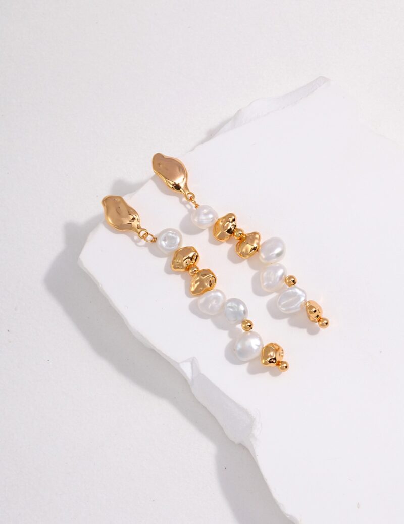 A pair of elegant earrings with gold clasps linked to a cascade of alternating freshwater pearls and small gold charms shaped like teddy bears, displayed on a textured white surface. Elegant pearl jewelry