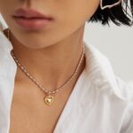 Close-up of a person wearing a white shirt and a heart-shaped gold pearl jewelry pendant, focusing on the lower half of the face and the necklace. Elegant pearl jewelry