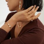 A woman with a bob haircut showcases elegant jewelry, including earrings, multiple rings, and bracelets adorned with freshwater pearls, against a neutral background. Her attire is a deep brown, complementing the. Elegant pearl jewelry