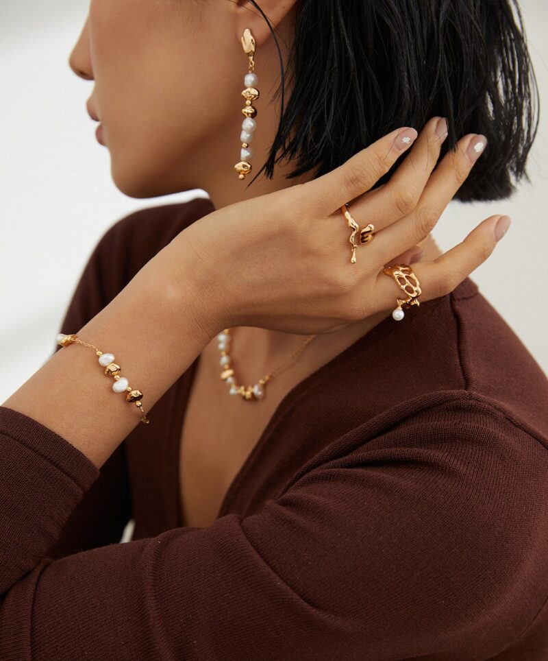 A woman with a bob haircut showcases elegant jewelry, including earrings, multiple rings, and bracelets adorned with freshwater pearls, against a neutral background. Her attire is a deep brown, complementing the. Elegant pearl jewelry