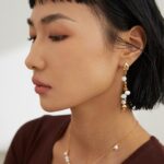 Profile view of a woman with a bob haircut showcasing elegant earrings and a matching necklace. She is wearing a brown top and has subtle makeup. Elegant pearl jewelry