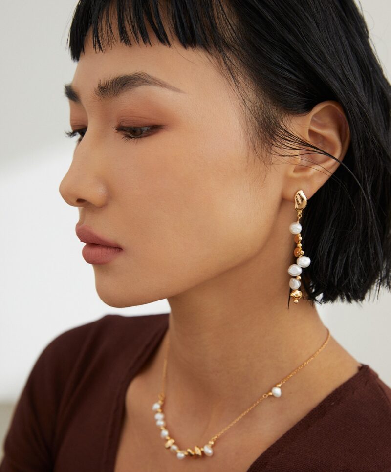 Profile view of a woman with a bob haircut showcasing elegant earrings and a matching necklace. She is wearing a brown top and has subtle makeup. Elegant pearl jewelry