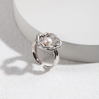 A pearl ring with an intricate floral design and a small freshwater pearl centerpiece, displayed atop a white circular stand against a textured grey background. Elegant pearl jewelry