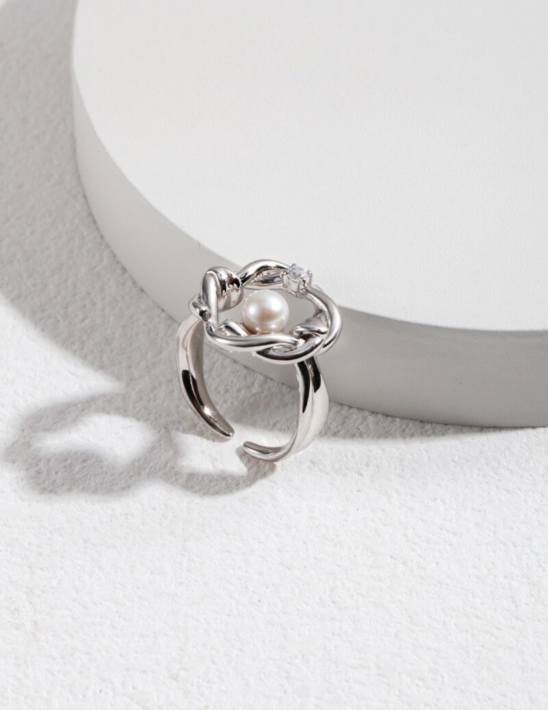 A pearl ring with an intricate floral design and a small freshwater pearl centerpiece, displayed atop a white circular stand against a textured grey background. Elegant pearl jewelry