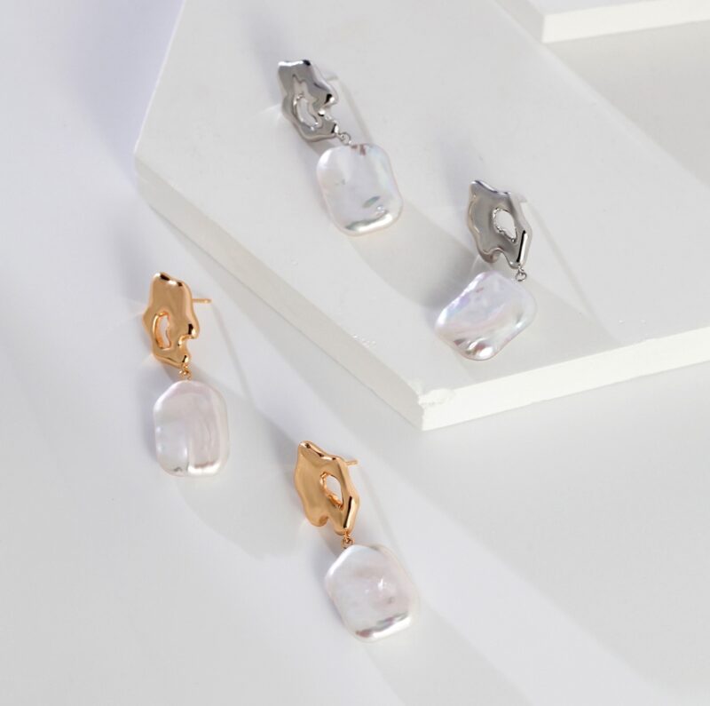 Three pairs of Elegant pearl jewelry with shimmering iridescent rectangular pendants hanging from gold and silver clasps, presented on a geometric white display stand adorned with freshwater pearl. Elegant pearl jewelry