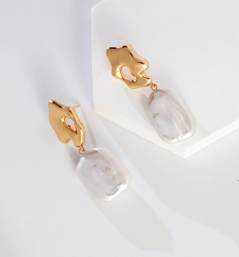 A pair of elegant earrings featuring golden abstract shapes connected to iridescent white pearl-like pendants, part of our jewelry collection, showcased on a white geometric stand with a soft shadow. Elegant pearl jewelry