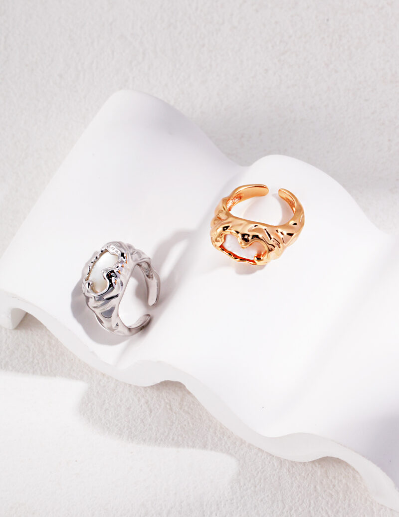 Two artistic rings displayed on a wavy white surface: one ring textured like a wave, and the other gold, resembling ornate, intertwined foliage adorned with a natural pearl. Elegant pearl jewelry