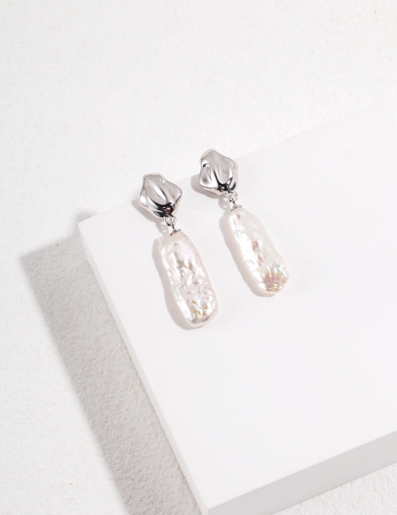 A pair of gold metallic tops and rectangular freshwater pearl pendants, displayed on a white surface with a subtle textured background. Elegant pearl jewelry