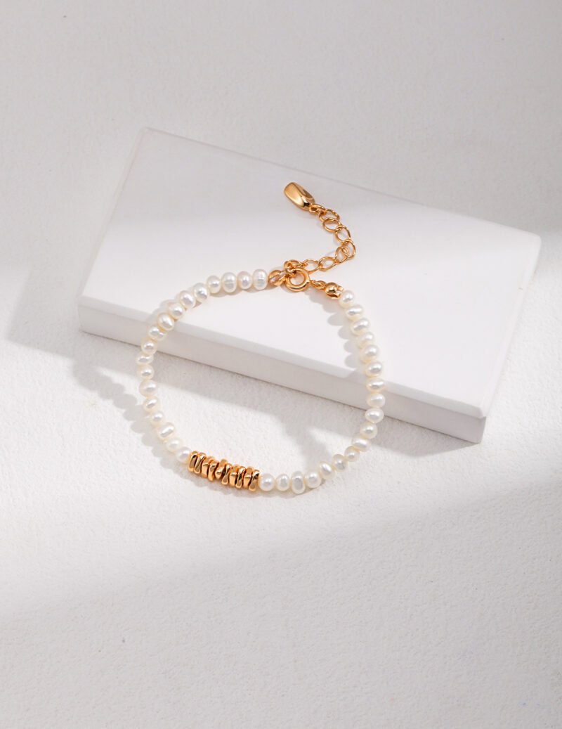 A bracelet with a gold clasp, categorized as gold pearl jewelry, rests on a white rectangular box placed on a light grey textured surface, illuminated by natural light. Elegant pearl jewelry