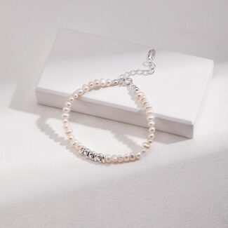 A bracelet with a metallic clasp lies atop a white rectangular box on a textured surface, illuminated by natural light casting soft shadows. Elegant pearl jewelry