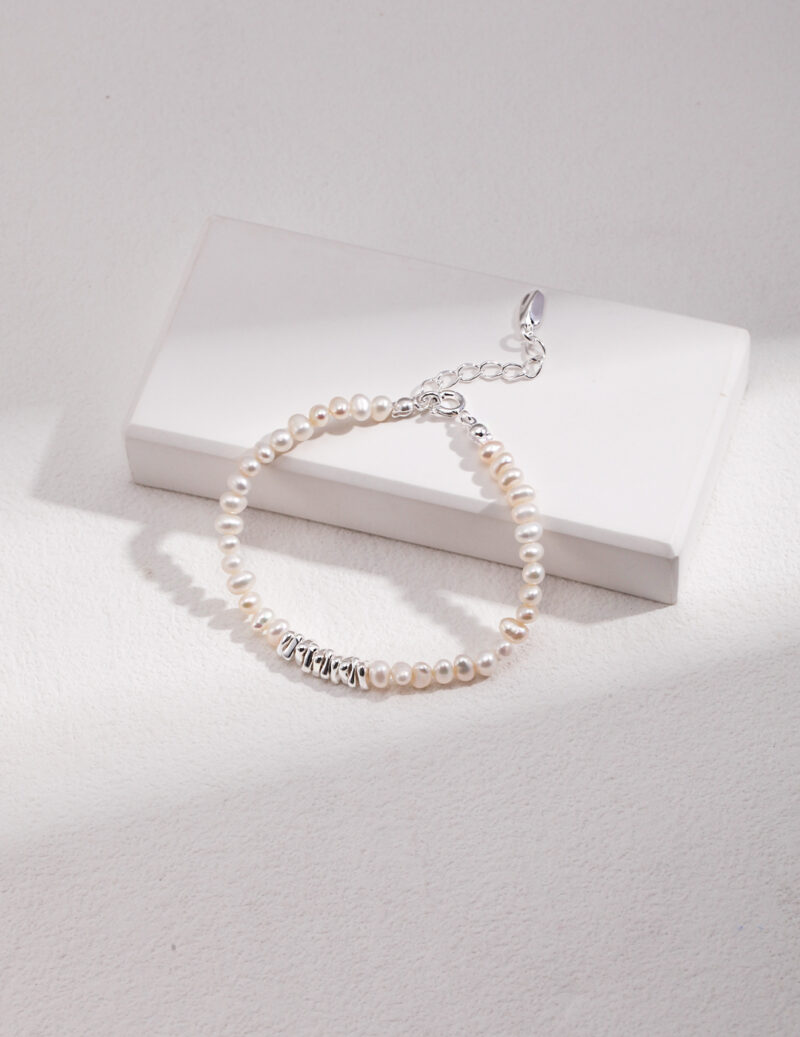 A bracelet with a metallic clasp lies atop a white rectangular box on a textured surface, illuminated by natural light casting soft shadows. Elegant pearl jewelry