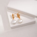 A pair of elegant gold earrings with natural pearl drops resting on a set against a textured beige background. Elegant pearl jewelry