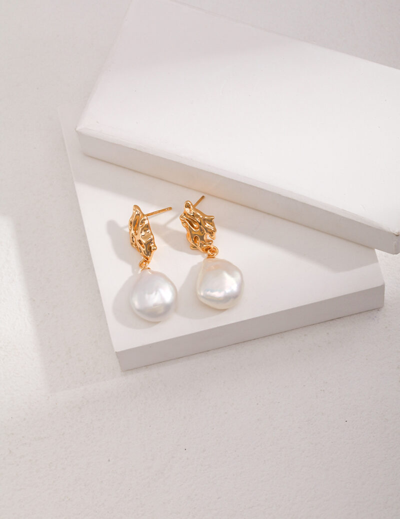 A pair of elegant gold earrings with natural pearl drops resting on a set against a textured beige background. Elegant pearl jewelry