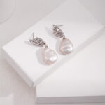 A pair of elegant earrings with intricate sterling details, displayed on a white stand against a soft-textured background. Elegant pearl jewelry