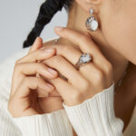 A close-up of a woman's hands clasped near her chin, showcasing elegant jewelry on her fingers. She wears a white blouse and a pearl earring, with a braided hairstyle. Elegant pearl jewelry