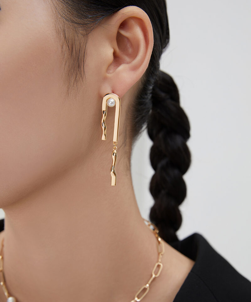 Profile view of a woman wearing unique dangling earring with a pearl accent and a matching gold necklace, against a light background. Her hair is styled in a neat braid Elegant pearl jewelry