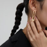 A woman with a braided ponytail wears a large pair of earring and multiple rings, touching her earring with her hand, against a grey background. Elegant pearl jewelry