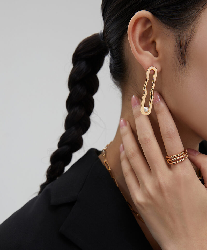 A woman with a braided ponytail wears a large pair of earring and multiple rings, touching her earring with her hand, against a grey background. Elegant pearl jewelry