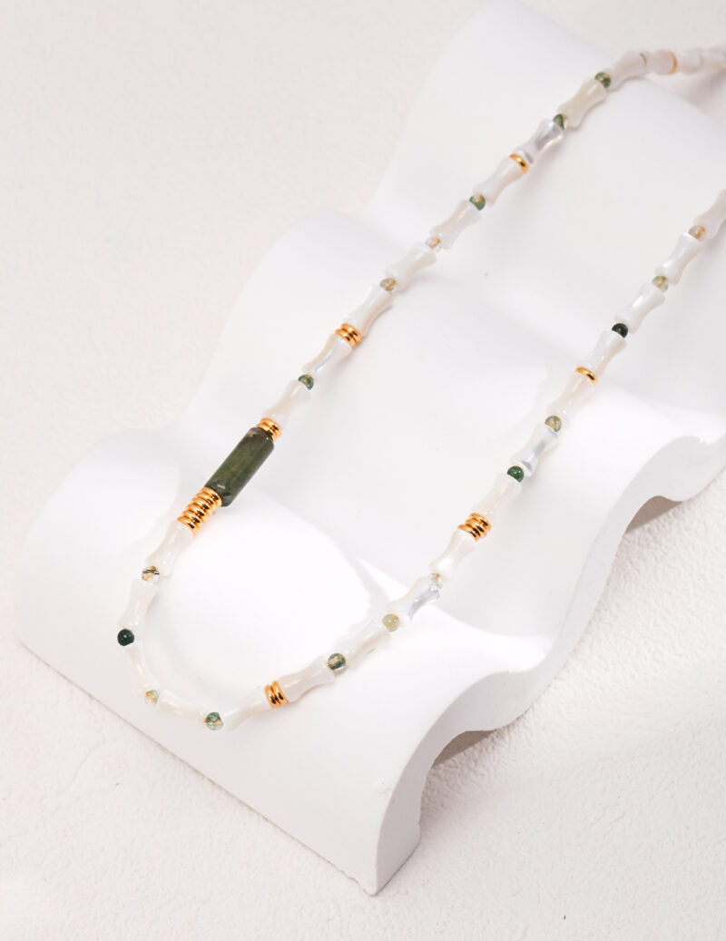 A delicate necklace with green and clear beads, highlighted by stone accents, draped gracefully on a white, wave-shaped jewelry stand against a cream background. Elegant pearl jewelry