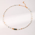 A delicate necklace featuring white and green beads with small gold accents and freshwater pearls, displayed on a white textured background. The clasp and chain are visible, hinting at a customizable length. Elegant pearl jewelry