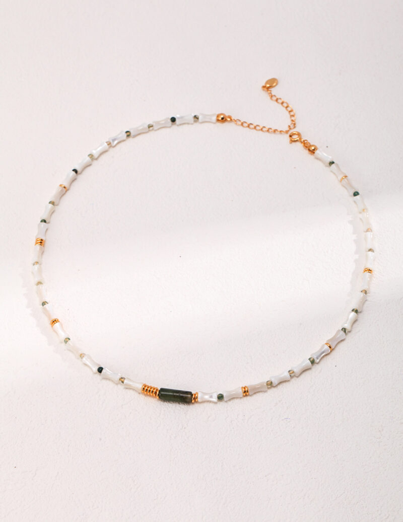 A delicate necklace featuring white and green beads with small gold accents and freshwater pearls, displayed on a white textured background. The clasp and chain are visible, hinting at a customizable length. Elegant pearl jewelry