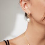 Close-up of a woman wearing an elegant earring with a large freshwater pearl and gold details, and a delicate gold necklace. Focus is on the earring and her profile. Elegant pearl jewelry