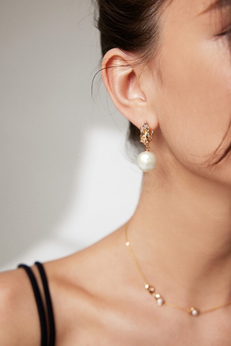 Close-up of a woman wearing an elegant earring with a large freshwater pearl and gold details, and a delicate gold necklace. Focus is on the earring and her profile. Elegant pearl jewelry