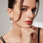 Close-up portrait of a woman with her hand on her chin, wearing a black top and elegant earrings, showcasing a subtle makeup look and an introspective gaze. Elegant pearl jewelry