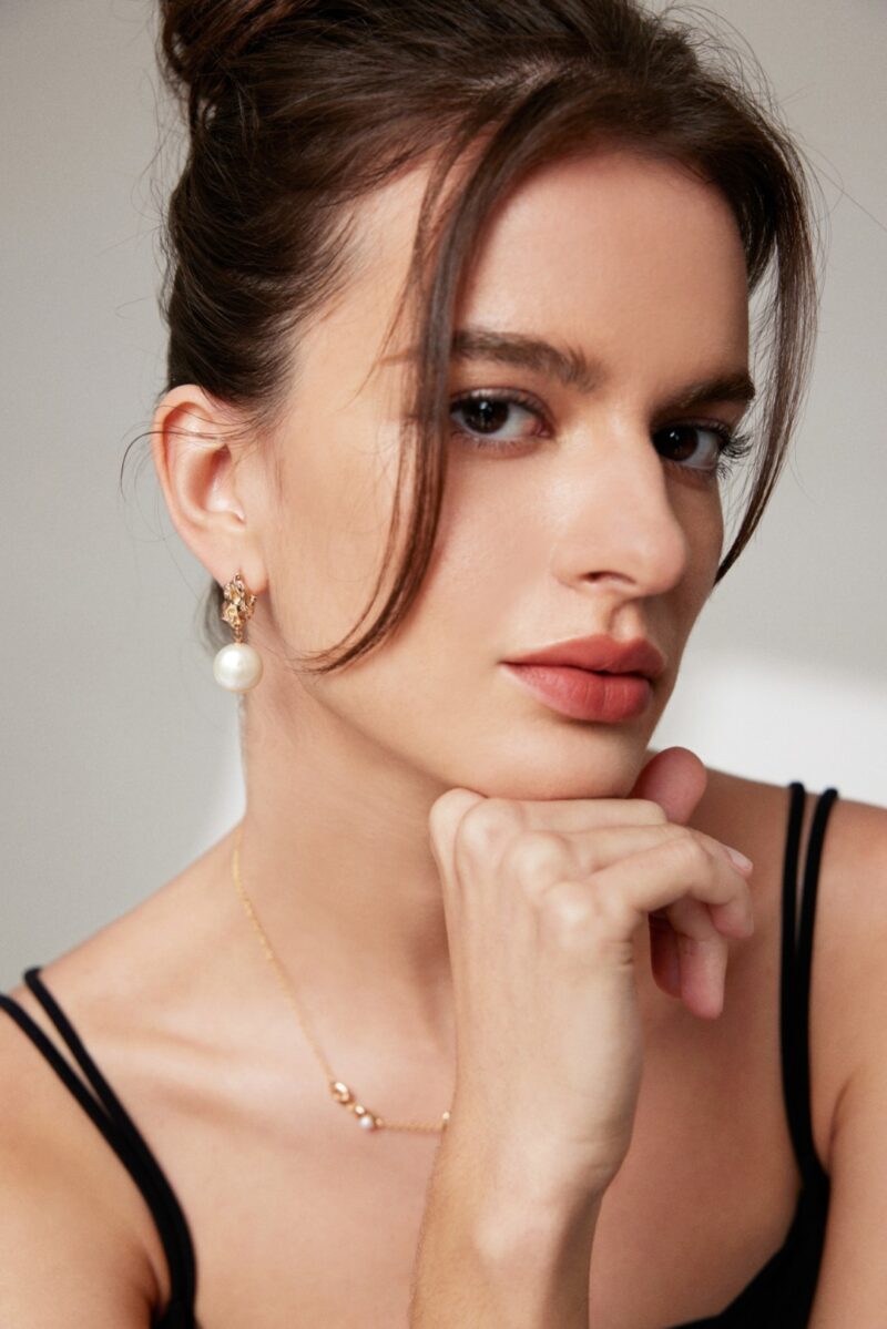 Close-up portrait of a woman with her hand on her chin, wearing a black top and elegant earrings, showcasing a subtle makeup look and an introspective gaze. Elegant pearl jewelry