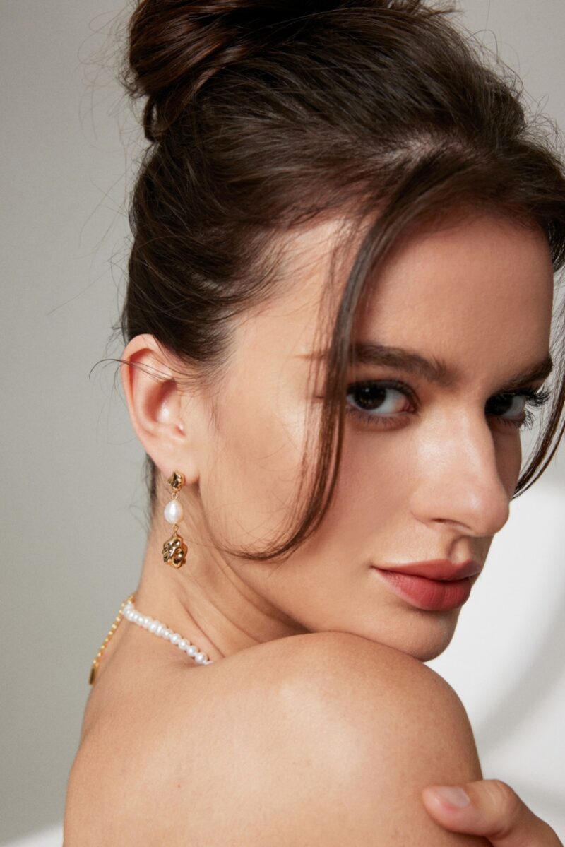 Close-up portrait of a woman with a bun hairstyle, wearing drop earrings and a pearl necklace, looking over her shoulder with a soft gaze. Elegant pearl jewelry