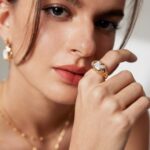 A close-up portrait of a woman with light makeup, partially covering her face with her hand adorned with a ring and matching pearl earrings. Elegant pearl jewelry