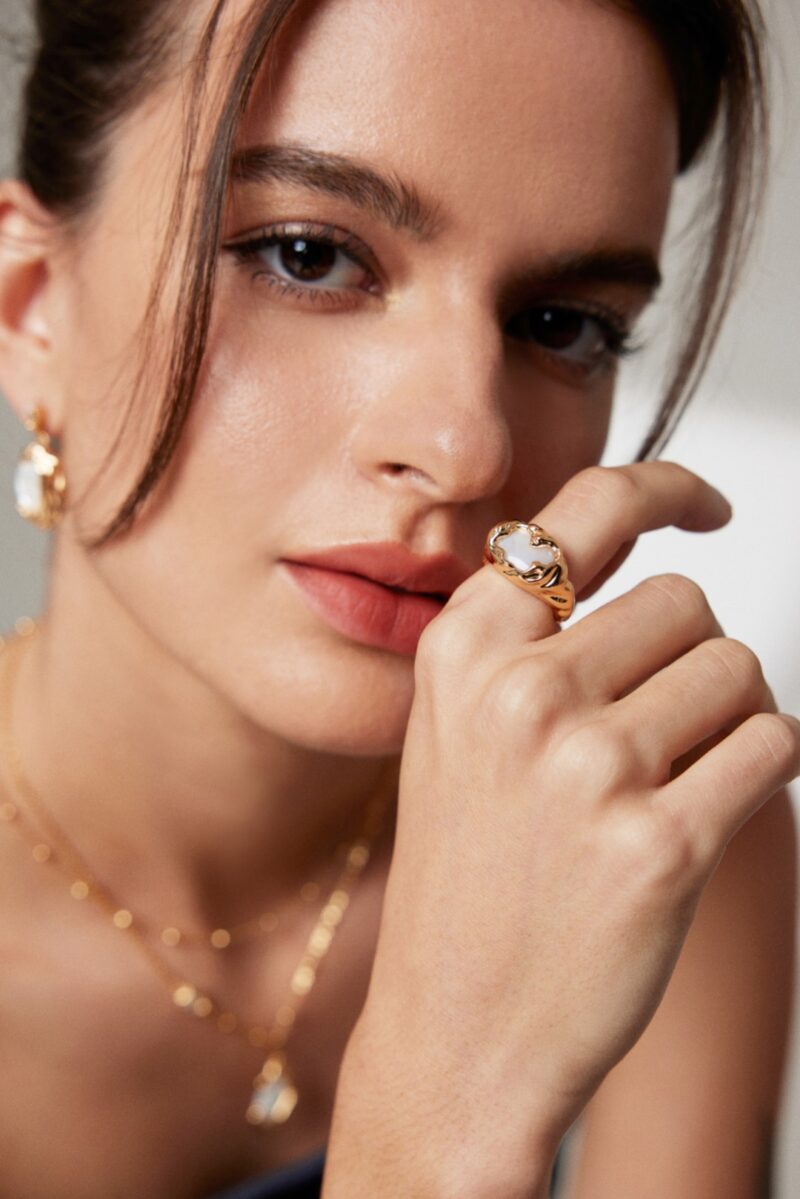 A close-up portrait of a woman with light makeup, partially covering her face with her hand adorned with a ring and matching pearl earrings. Elegant pearl jewelry