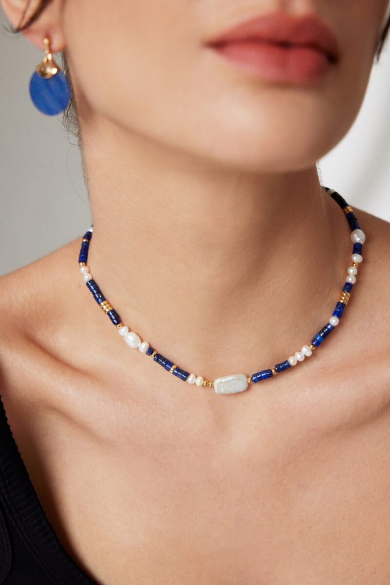 Sentence with product name: Close-up of a woman wearing a necklace featuring a central natural pearl pendant, paired with a matching blue earring. Elegant pearl jewelry