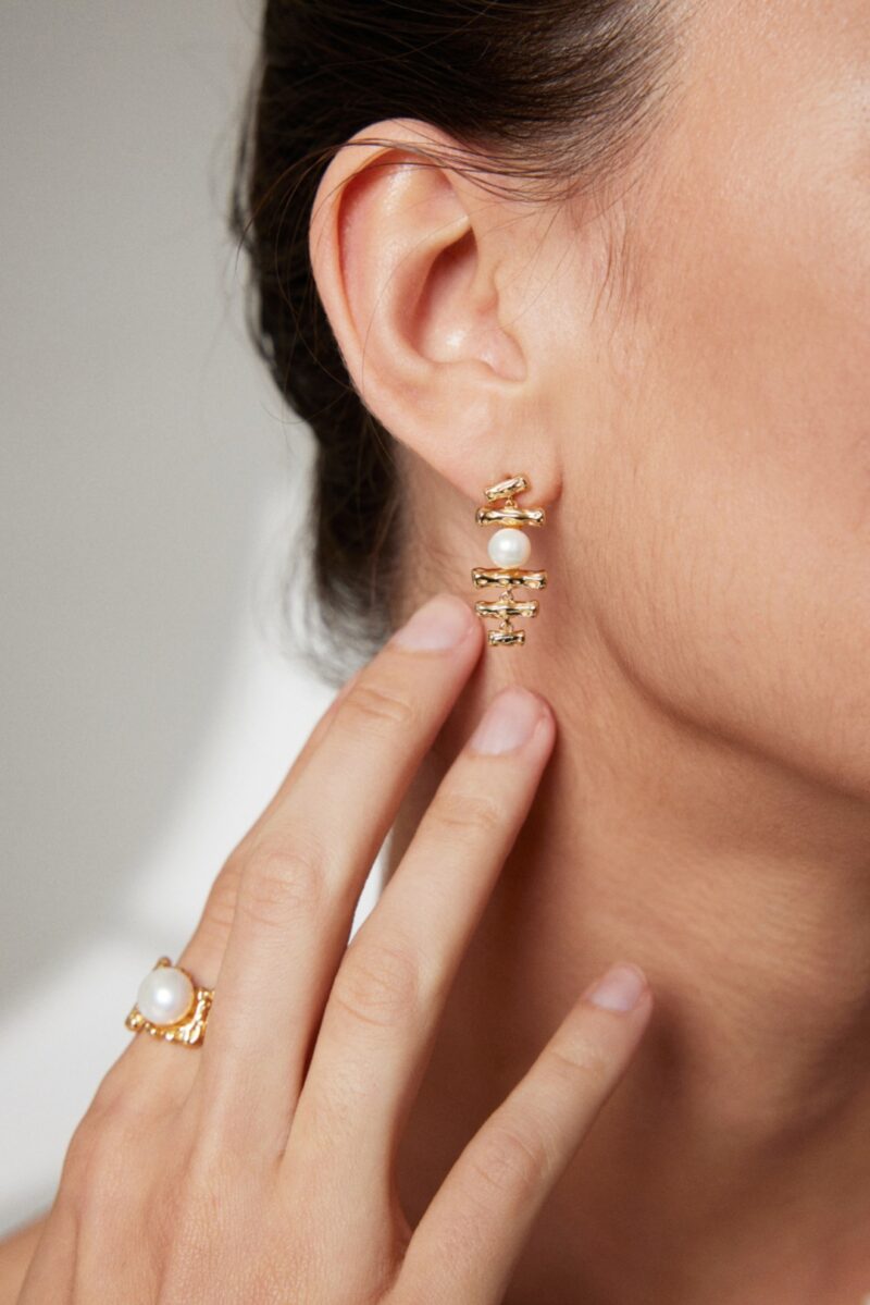 Close-up view of a woman's ear wearing an elegant earring, with her hand showcasing a matching ring. Her smooth skin and refined jewelry highlight a sophisticated style. Elegant pearl jewelry