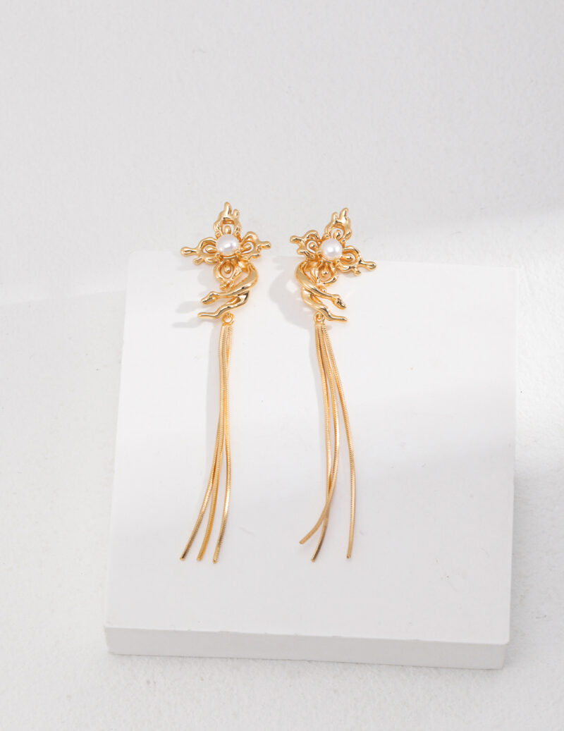 A pair of intricate  tassel earrings displayed on a plain white stand against a light gray background. The earrings have a detailed floral design at the top featuring natural pearl. Elegant pearl jewelry