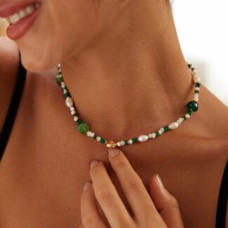 A woman wearing a black top is shown smiling, adorned with the o_1i3hbdovv1k6q8q91f9a1n0l9j13 pearl and malachite necklace, her finger gently touching it. She also wears gold-framed, square-shaped pearl earrings. The background is softly blurred. Elegant pearl jewelry