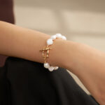 A close-up image of a woman's wrist adorned with a o_1fo4sbs30nu015bose7vqa5ais bracelet featuring a gold clasp, set against a backdrop of her maroon and black outfit. Elegant pearl jewelry