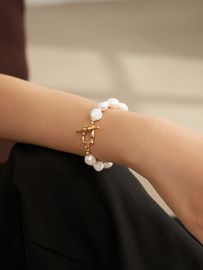 A close-up image of a woman's wrist adorned with a o_1fo4sbs30nu015bose7vqa5ais bracelet featuring a gold clasp, set against a backdrop of her maroon and black outfit. Elegant pearl jewelry