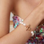 A close-up of a woman's wrist adorned with a o_1hkinrhen1ofjllos621dar1j1s0 bracelet featuring an 18k gold toggle clasp, set against a sparkling multicolored dress and a pearl necklace. Elegant pearl jewelry