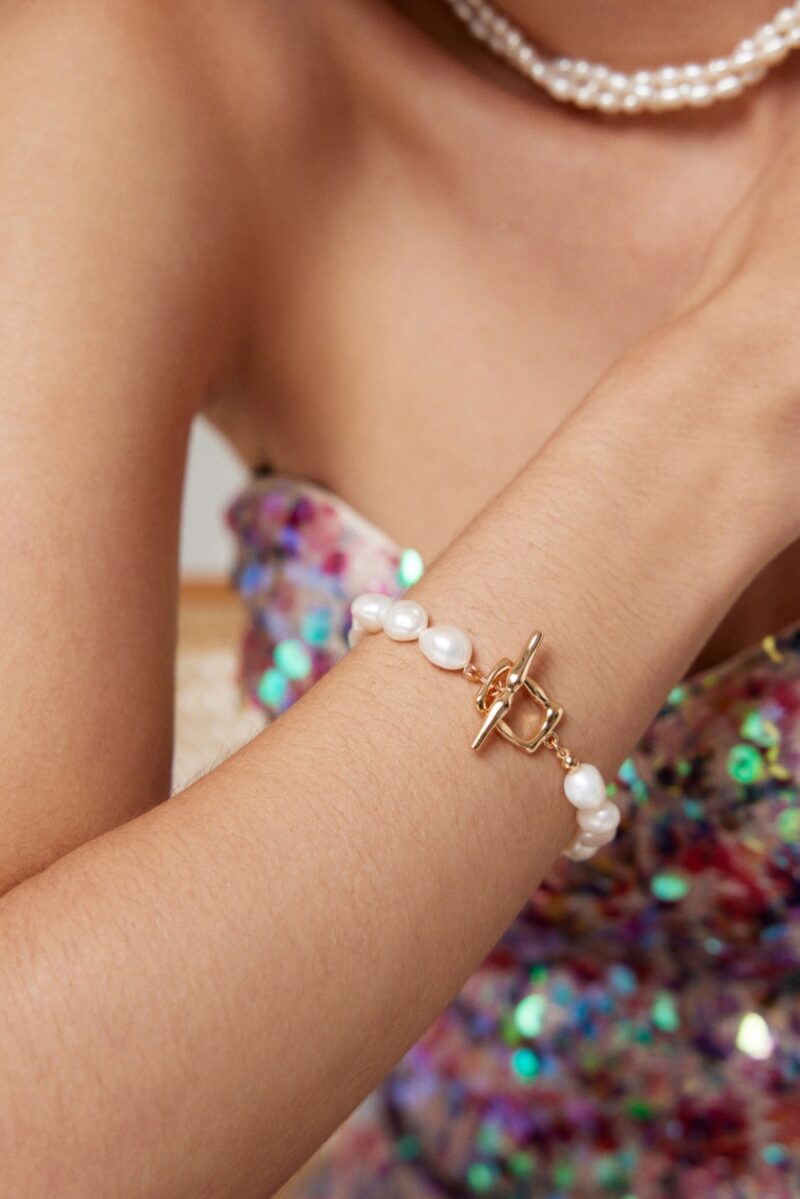 A close-up of a woman's wrist adorned with a o_1hkinrhen1ofjllos621dar1j1s0 bracelet featuring an 18k gold toggle clasp, set against a sparkling multicolored dress and a pearl necklace. Elegant pearl jewelry