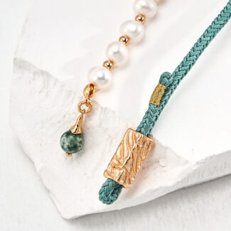 A stylish o_1ht8oi7td1ln31seg7111ajb1ae91 displayed on a white textured surface, featuring a combination of pearl beads, a gold vermeil pendant, and a green braided cord with a Jasper stone charm. Elegant pearl jewelry