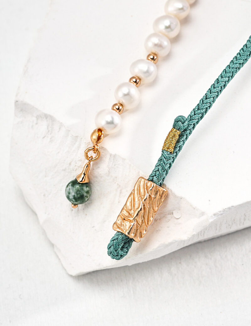 A stylish o_1ht8oi7td1ln31seg7111ajb1ae91 displayed on a white textured surface, featuring a combination of pearl beads, a gold vermeil pendant, and a green braided cord with a Jasper stone charm. Elegant pearl jewelry