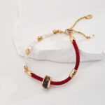 A red o_1hte7up811s4iggt1iqbgc01aoff braided bracelet with gold vermeil clasps and details, featuring a black gemstone and white pearls, displayed on a textured white stone surface. Elegant pearl jewelry