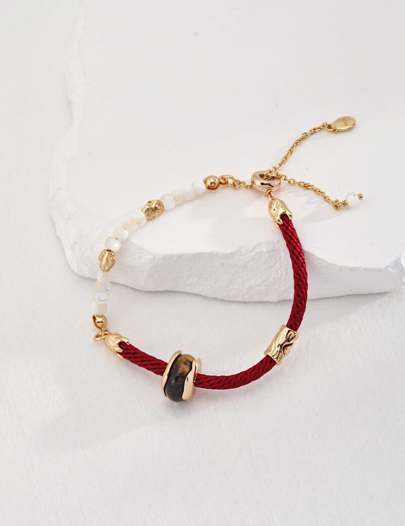 A red o_1hte7up811s4iggt1iqbgc01aoff braided bracelet with gold vermeil clasps and details, featuring a black gemstone and white pearls, displayed on a textured white stone surface. Elegant pearl jewelry