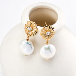 Gold earrings with intricate floral designs and embedded diamonds, part of our o_1htim9ik313mmbie1a0gq6p1u302, adorned with large, white, irregularly shaped pearls. Elegantly displayed hanging from the rim of a minimalist white ceramic vase, these 18k gold pearl earrings exude timeless elegance. Elegant pearl jewelry