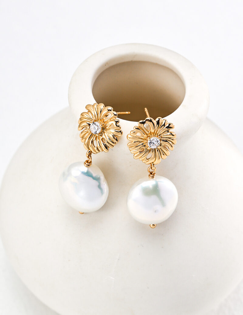 Gold earrings with intricate floral designs and embedded diamonds, part of our o_1htim9ik313mmbie1a0gq6p1u302, adorned with large, white, irregularly shaped pearls. Elegantly displayed hanging from the rim of a minimalist white ceramic vase, these 18k gold pearl earrings exude timeless elegance. Elegant pearl jewelry