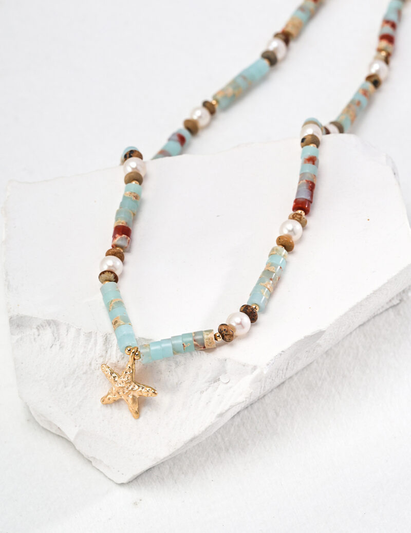 A necklace with beads in turquoise, white, tan, and rust colors, featuring a starfish pendant in gold, is displayed on a broken white ceramic piece. The o_1hucrll2csj615l1ap51arbpdic is casually draped over the ceramic, highlighting its coastal and bohemian style—perfect for some summer fun. Elegant pearl jewelry