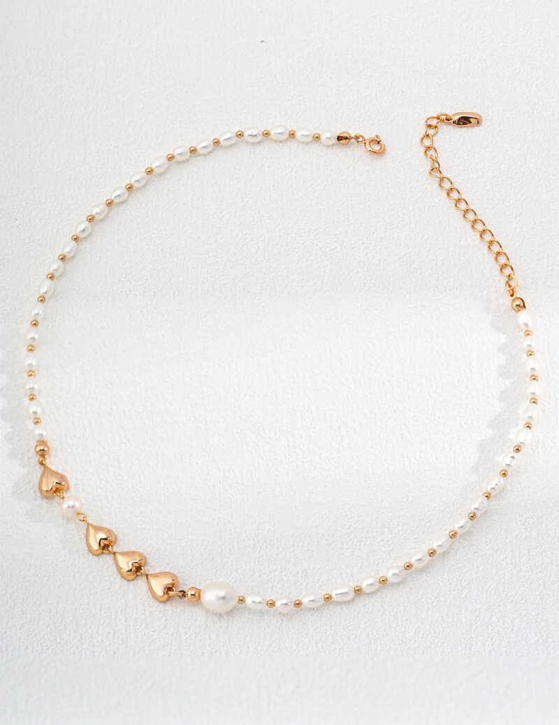 A delicate o_1hv8s3jhrkjha2o1gqs1hku1rsm3 with small gold heart accents is laid out on a white surface. The necklace features an adjustable gold chain clasp and is adorned with evenly spaced pearls and heart-shaped gold pieces for an elegant, minimalist design. Elegant pearl jewelry