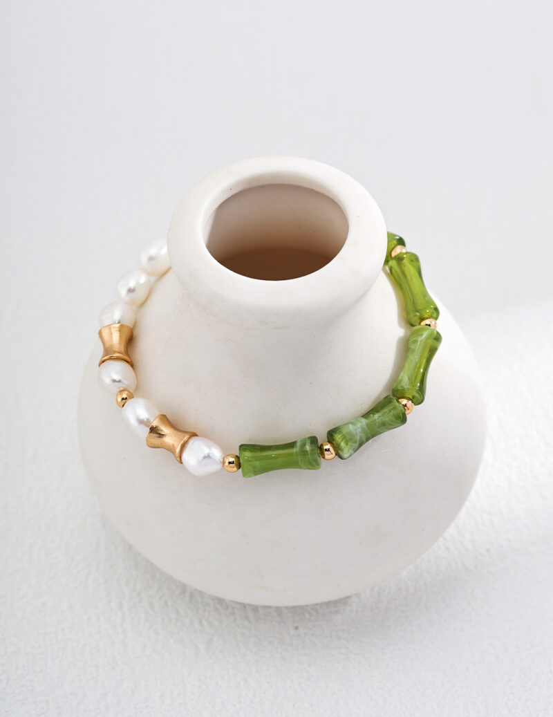 A o_1i0aipujd18u8no72v71fbd1has3 featuring alternating white pearls, 18k gold vermeil star-shaped beads, and green cylindrical beads is draped around the neck of a small, white ceramic vase with a smooth finish. The vase sits against a soft white background. Elegant pearl jewelry