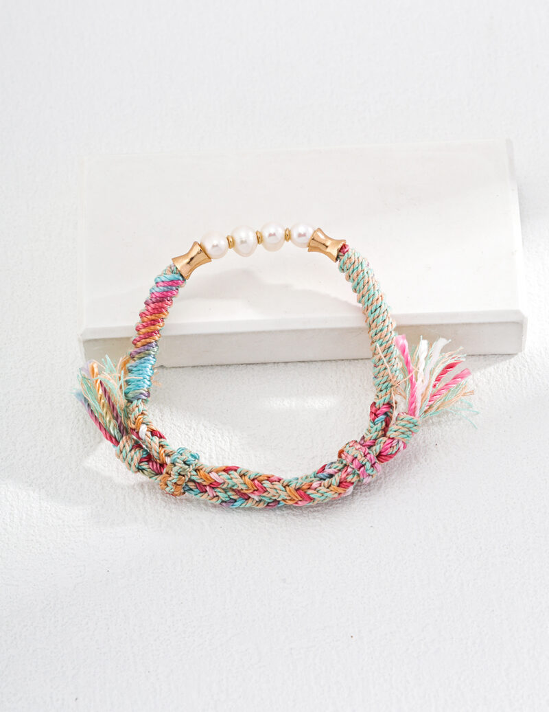 A colorful braided friendship bracelet with a mix of pastel threads and small tassels on each end, adorned with five white beads in the center, displayed on a white background. Exuding rustic elegance, the o_1i0d4pcs51nup1uluo5fvoj1of95 rests on a rectangular white platform beneath part of the bracelet. Elegant pearl jewelry